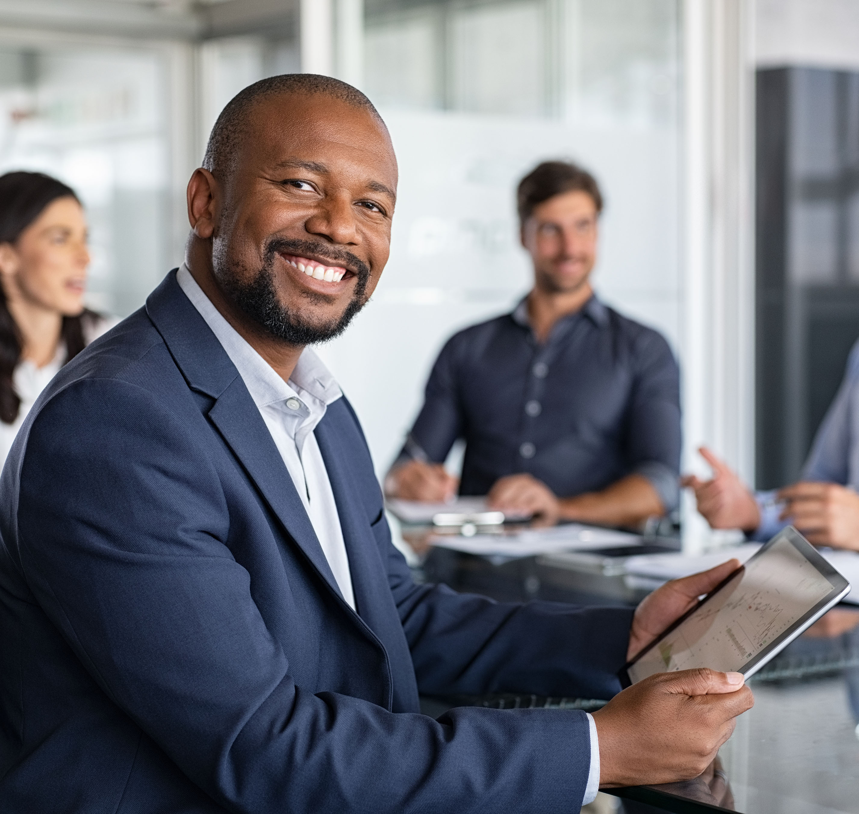 Mature black businessman with colleagues sitting in a modern board room. Proud smiling business man sitting during a meeting and looking at camera. Portrait of happy successful executive with team working in background.